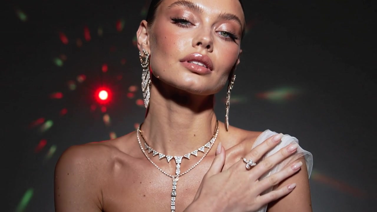 Modella in posa in studio, che si tocca il petto e inclina la testa sotto una luce direzionata, mostrando una collana a strati e un drappeggio.