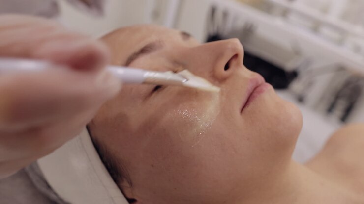 Kosmetikerin, die im Rahmen einer Enzymtherapie mit Pinseln eine feuchtigkeitsspendende medizinische Creme auf das Gesicht einer jungen Frau aufträgt