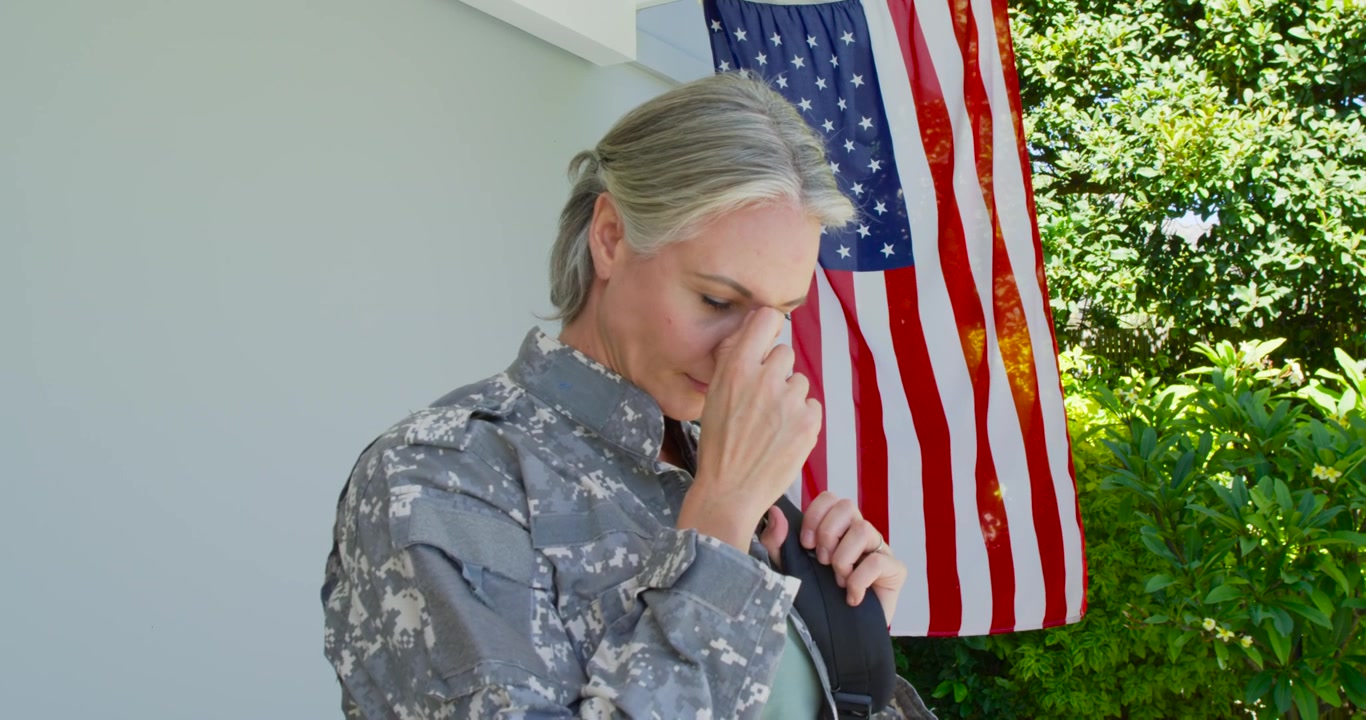 Donna di mezza età in mimetica che sistema la cinghia dello zaino, si strofina il naso riflettendo sul portico accanto alla bandiera americana.