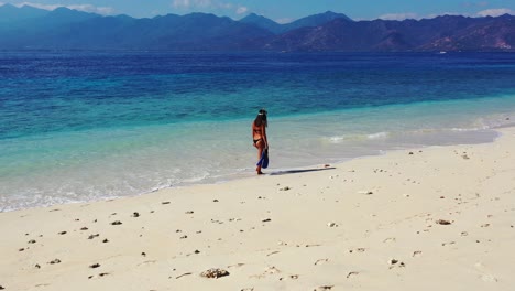 Woman-walking-through-the-shallow-ocean-waves-by-herself-down-a-beautiful-beach-shoreline