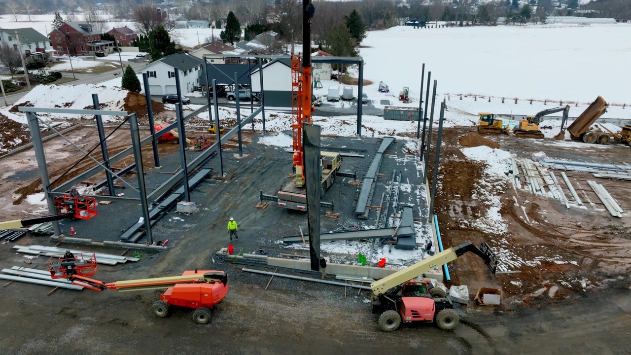 Construction is in progress at a building site during winter. Workers operate machinery to build a structure. Cranes lift materials and the site is covered with snow.