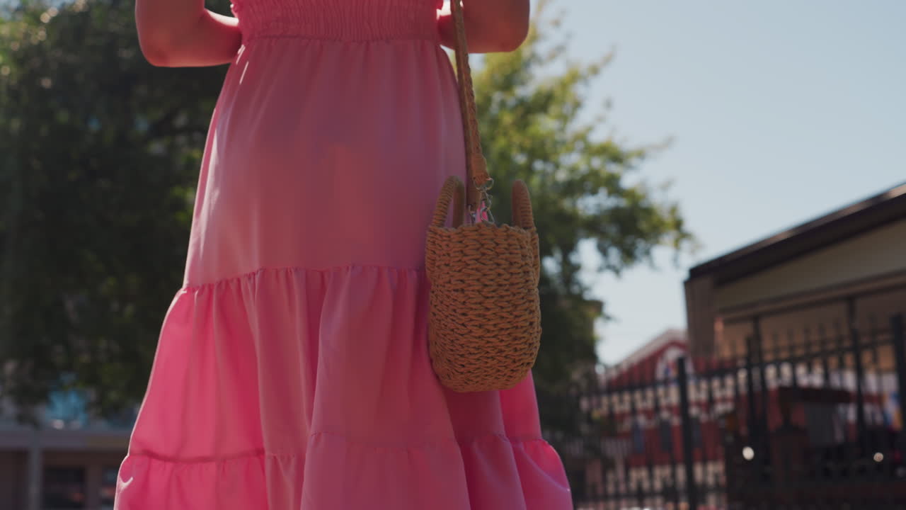 Donna con abito rosa e borsa di paglia, primo piano attraverso un giardino cittadino illuminato dal sole, dettaglio della borsa intrecciata e gonna a balze fluente, sfondo bokeh delicato, brezza leggera, atmosfera da mercato estivo, stile da fashion blogger
