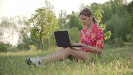 woman freelancer working remotely sitting on grass in public park using laptop.