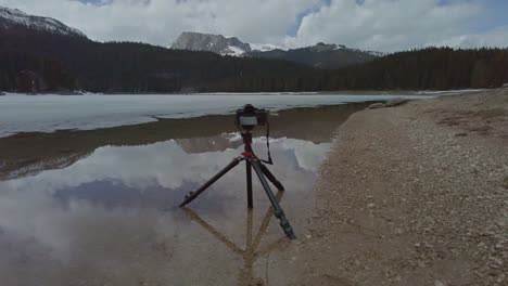 a camera on a tripod sitting in lake crno jezero, albania taking landscape photos of snow-capped mountains