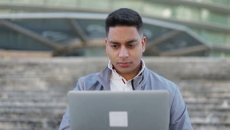 Un-Joven-Enfocado-Usando-Una-Computadora-Portátil-Mientras-Está-Sentado-Al-Aire-Libre