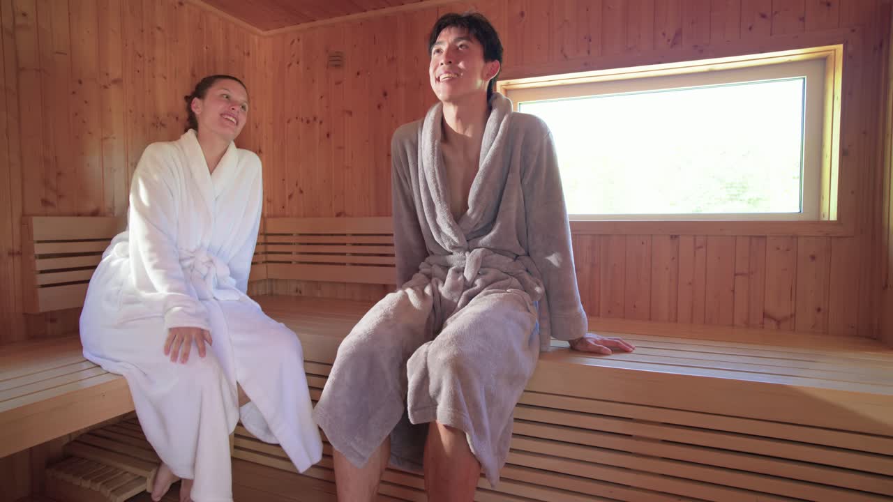 Coppia che entra in sauna, l'uomo si siede, la donna si unisce, chiacchierano e si rilassano sulla panca in accappatoio.