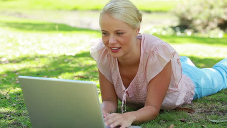 a woman lying in the park with her laptop as she then looks at the camera