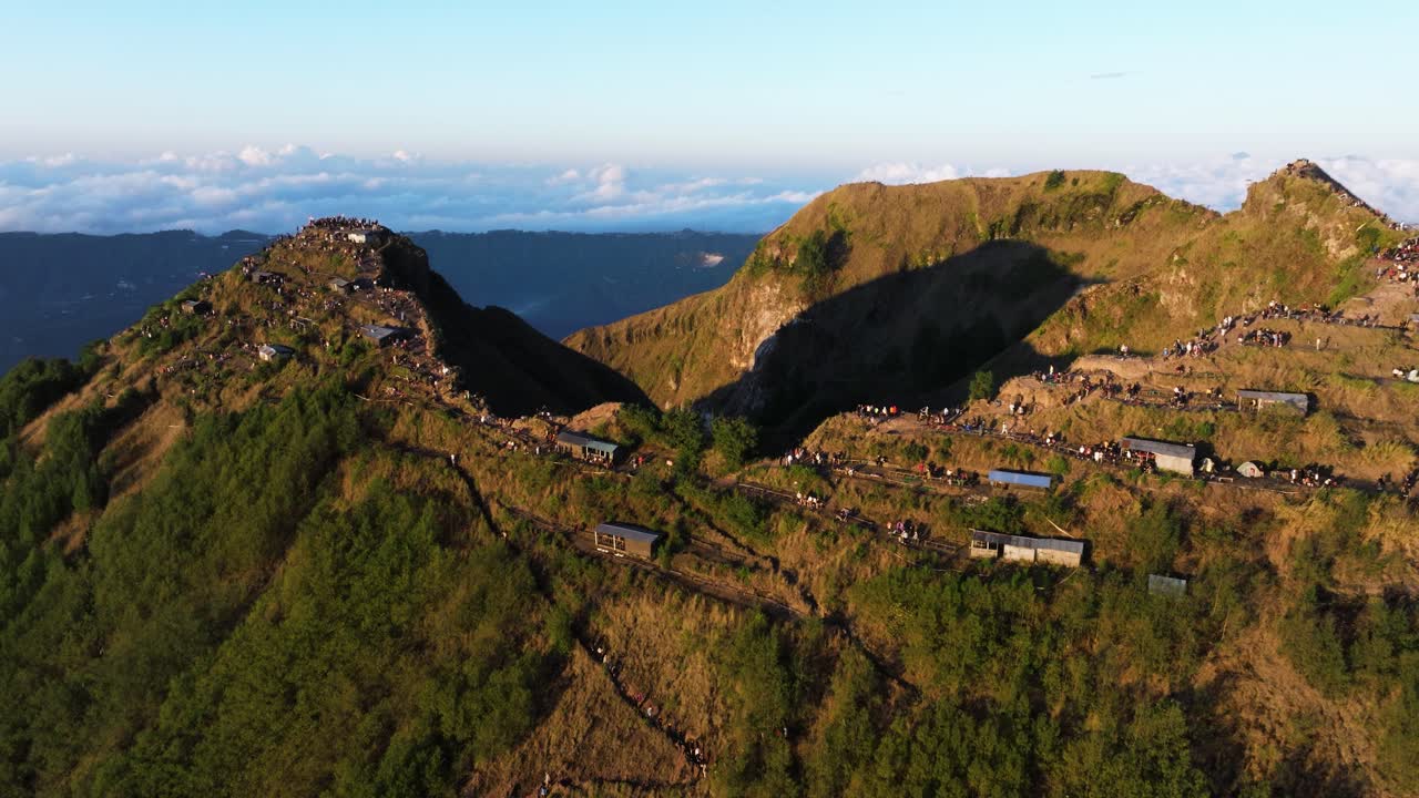 Sunlit Mount Batur summit with hikers on trails, surrounded by green volcanic landscape, aerial dolly to crowds along cliff edge