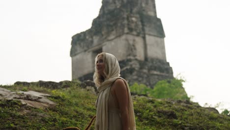 a woman draped in a cream scarf admires a towering mayan pyramid in the lush jungles of tikal, guatemala.