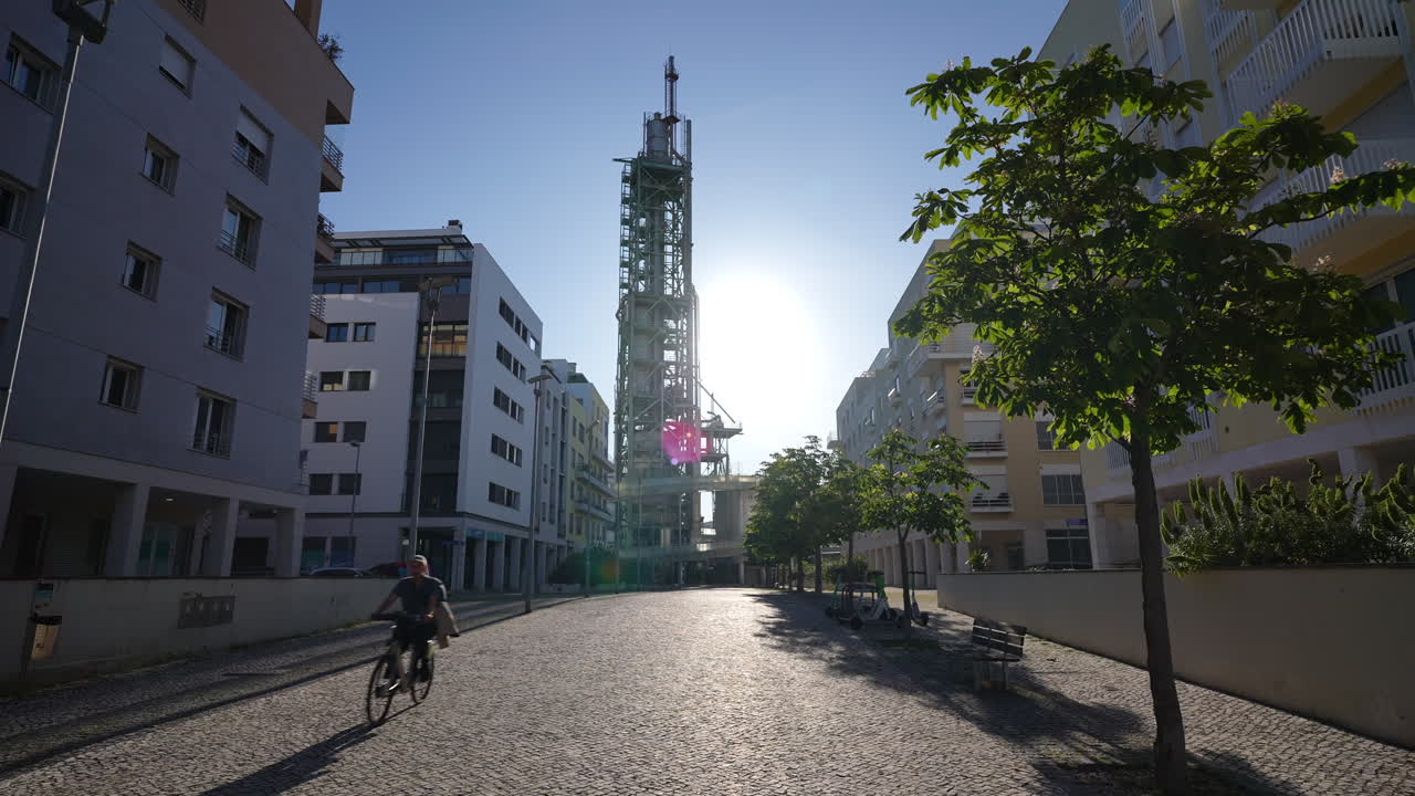 Urban street scene featuring modern buildings and a towering industrial structure bathed in sunlight, creating a striking contrast between nature and architecture in a vibrant city environment