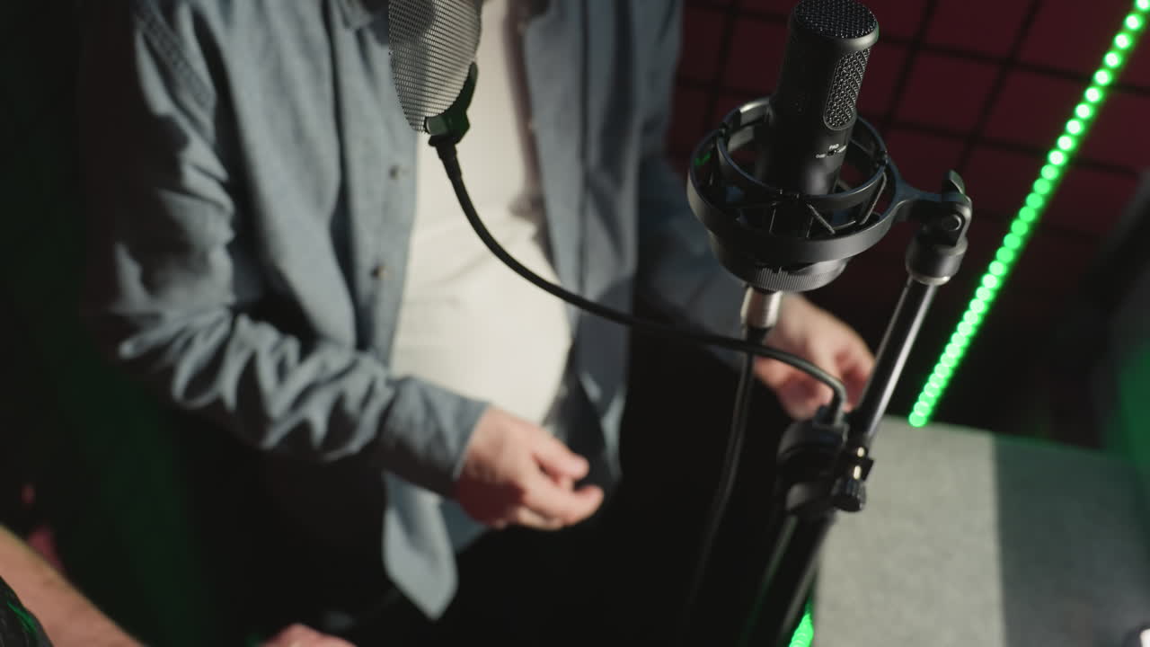 스튜디오 마이크의 클로즈업과 얼굴이 보이지 않는, 파란색 셔츠와 색 하 셔츠를 입은 남자가 음향 방역 녹음 스튜디오에서 손가락을 리적으로 니다.