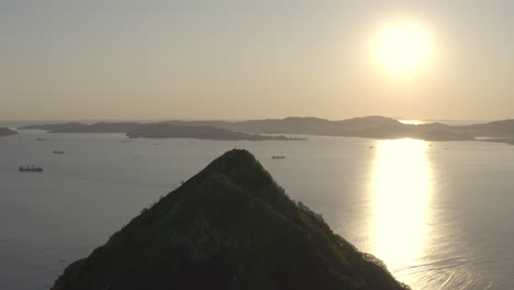 Una-Cumbre-De-Una-Montaña-En-Forma-De-Pirámide-Con-La-Bahía-Y-Los-Barcos-En-El-Fondeadero-En-El-Fondo,-Y-Los-Soles-Proyectando-Un-Gran-Reflejo-De-Rayos-En-La-Superficie-Del-Agua,-En-La-Puesta-De-Sol