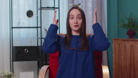 excited amazed young woman raising hands in surprise shocked by sudden victory at home on chair