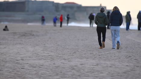 Dos-Mujeres-Caminan-Por-La-Playa-Durante-El-Campeonato-De-Surf-De-Billabong,-Llenas-De-Gente-En-La-Playa-Con-Olas-Grandes-Y-Fuertes-Detrás,-Playa-De-Carcavelos,-Cascais,-Portugal