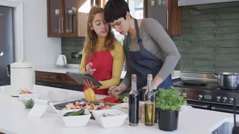 happy caucasian lesbian couple preparing food and using tablet in sunny kitchen