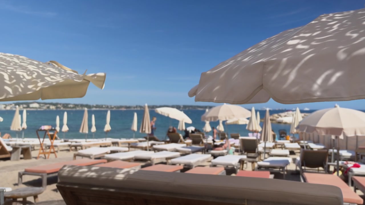 Area lounge di lusso sulla spiaggia con lettini, ombrelloni, mare calmo e yacht sullo sfondo.