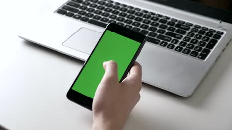 men's hands hold a big black smartphone scrolling gesture. green screen, chromakey concept, laptop in the background 60 fps