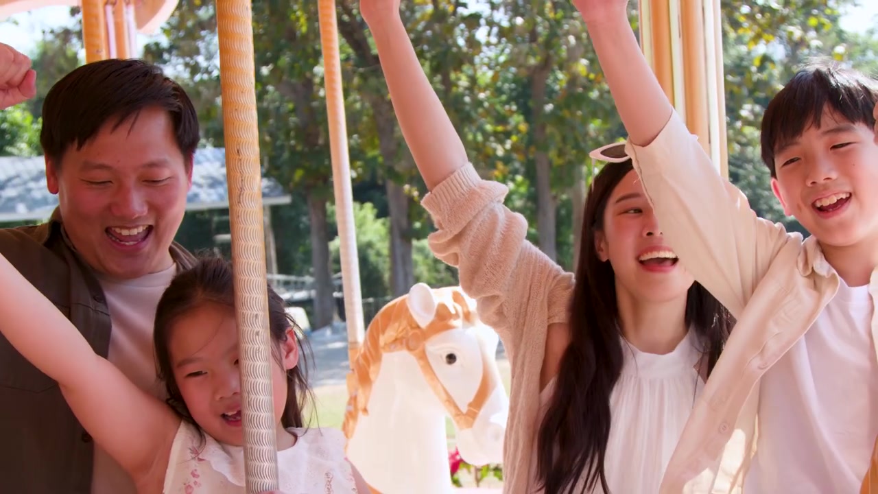 ครอบครัวชาวเอเชียที่ร่าเริงกำลังหัวเราะและโบกมือขณะเล่นม้าหมุนในเวลากลางวันที่สดใส