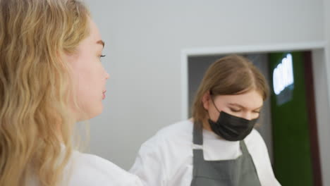 blonde-haired client with nose piercing examines face in handheld mirror after receiving eyebrow treatment from beautician wearing mask and apron in well-lit indoor salon environment