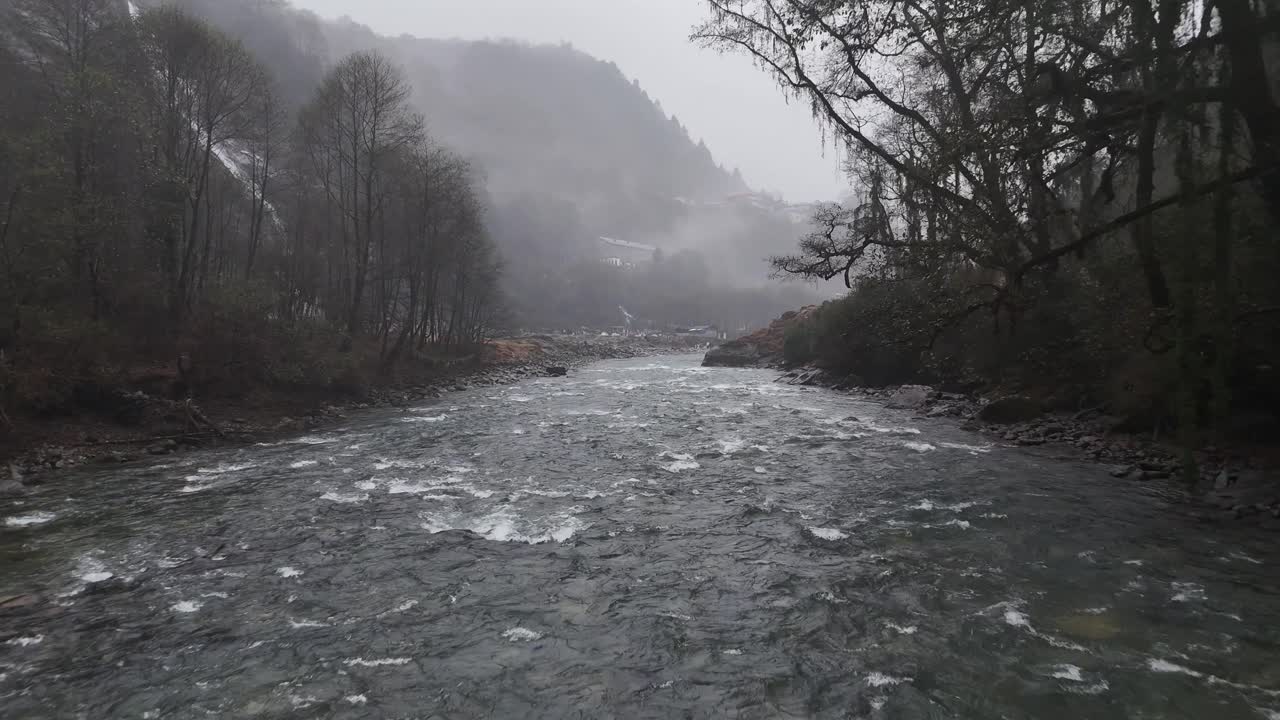 Hint Himalaya'larında, kayalık kıyılar ve çıplak kış ağaçlarıyla sisli ormanlık bir vadiden akan turkuaz renkli bir dağ nehrini atmosferik sis ve buğu içinde gösteren havadan drone görüntüleri.
