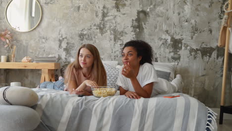 two female friends watching a movie at home. black girl and caucasian young woman eating popcorn lying on the bed.