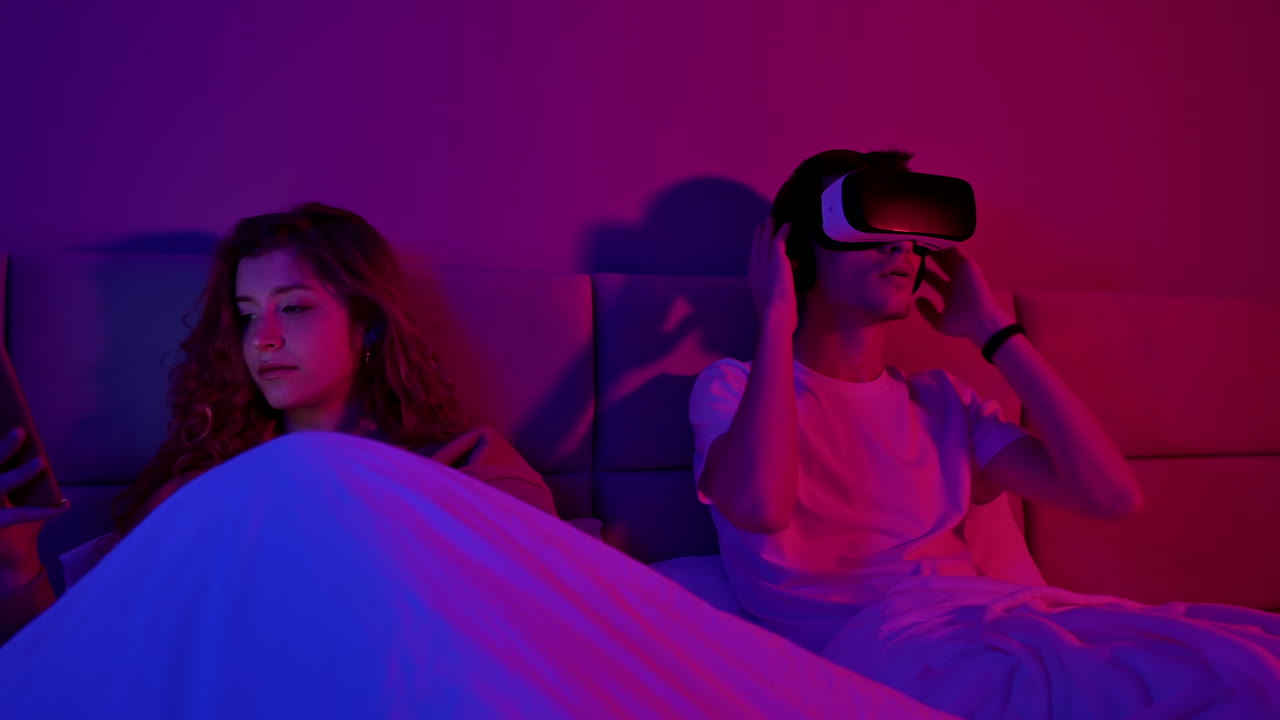 Young couple in the bedroom at night before sleeping. Man using virtual reality headset, girl browsing smartphone. Colorful lights