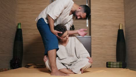 남성 태국 마사지사가 젊은 여성의 목을 팔꿈치 근처에 서서 마사지하고 있습니다.