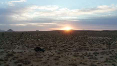 A-jeep-drives-along-a-dusty,-remote-trail-in-the-Mojave-Desert-with-the-sun-setting-beyond-the-horizon---aerial-view