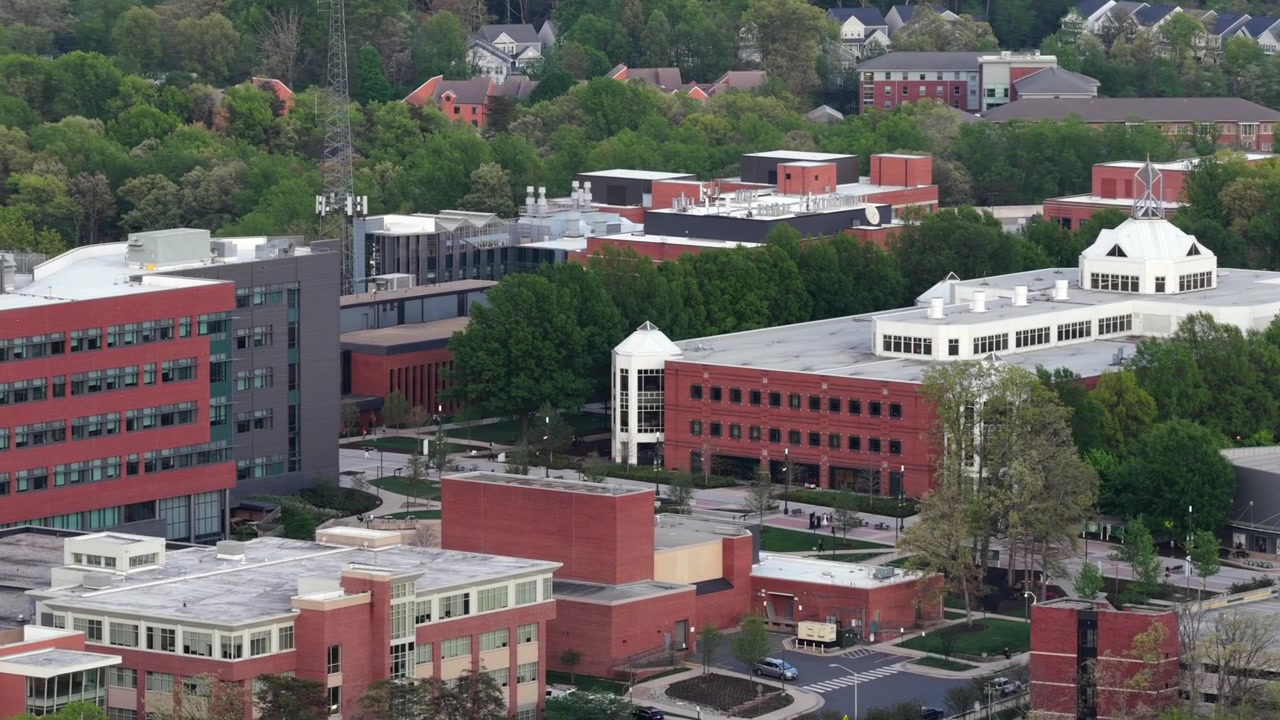 Veduta aerea del campus della George Mason University in Virginia, con moderni edifici accademici in mattoni, aree verdi alberate e un contesto suburbano. Scena iconica di un'istituzione educativa negli Stati Uniti. Inquadratura ampia.