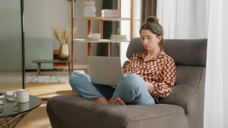 european lady sits cross-legged on lounge chair typing on laptop on lap