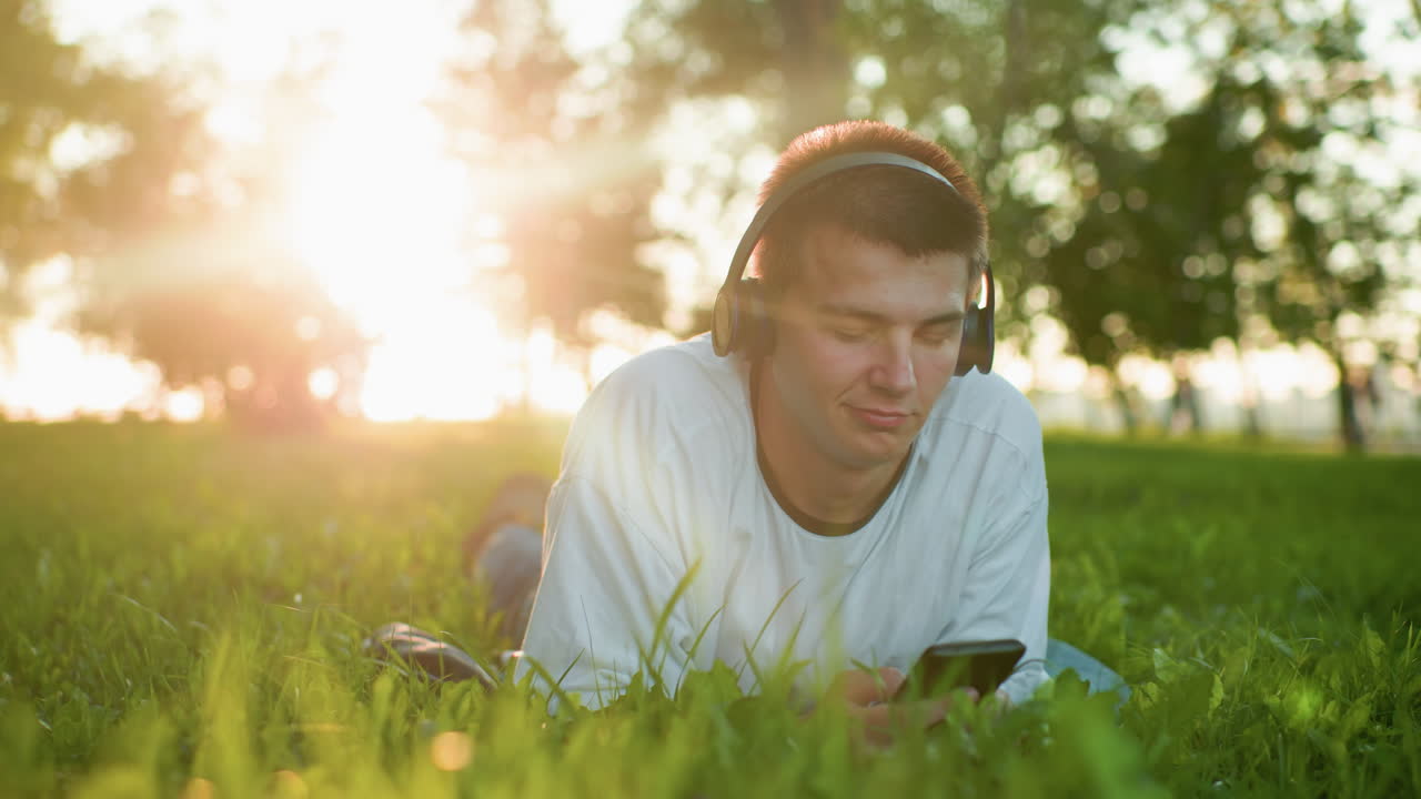 artista de música acostado en la hierba con auriculares disfrutando de la música mientras usa el teléfono, asintiendo con la cabeza al ritmo de la luz solar creando un efecto brillante en su cabeza, personas borrosas caminando en el fondo lejano