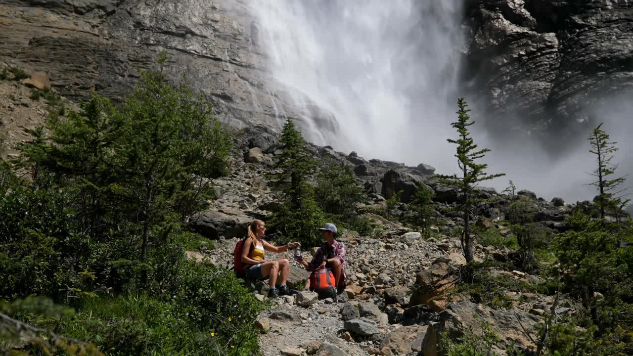 vista frontal de una joven pareja de excursionistas caucásicos con mochila sentados y relajándose en el bosque 4k