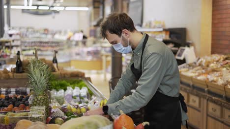 male assistant in supermarket food store worker in medical mask and apron arranges bananas on shelves