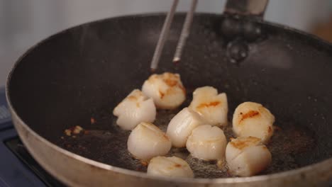 Vieiras-Cocinando-Y-Chisporroteando-En-Una-Sartén-Con-Aceite-Estallando-Y-Salpicando,-Chef-Volteando-Los-Lados-Con-Un-Par-De-Pinzas,-Tirando-Y-Girando-La-Sartén,-Escena-Culinaria-Llena-De-Humos-Aromáticos