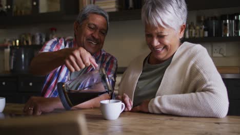Happy-senior-mixed-race-couple-pouring-coffee-laughing-in-kitchen