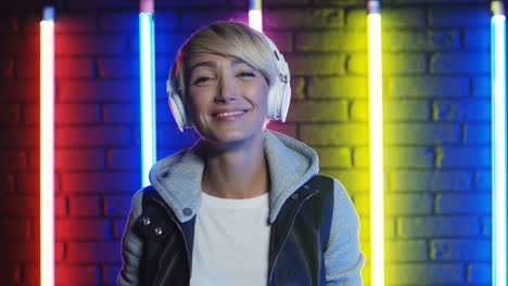 close up view of blonde woman putting on big white headphones and listening to music in a room with colorful neon lamps on the wall