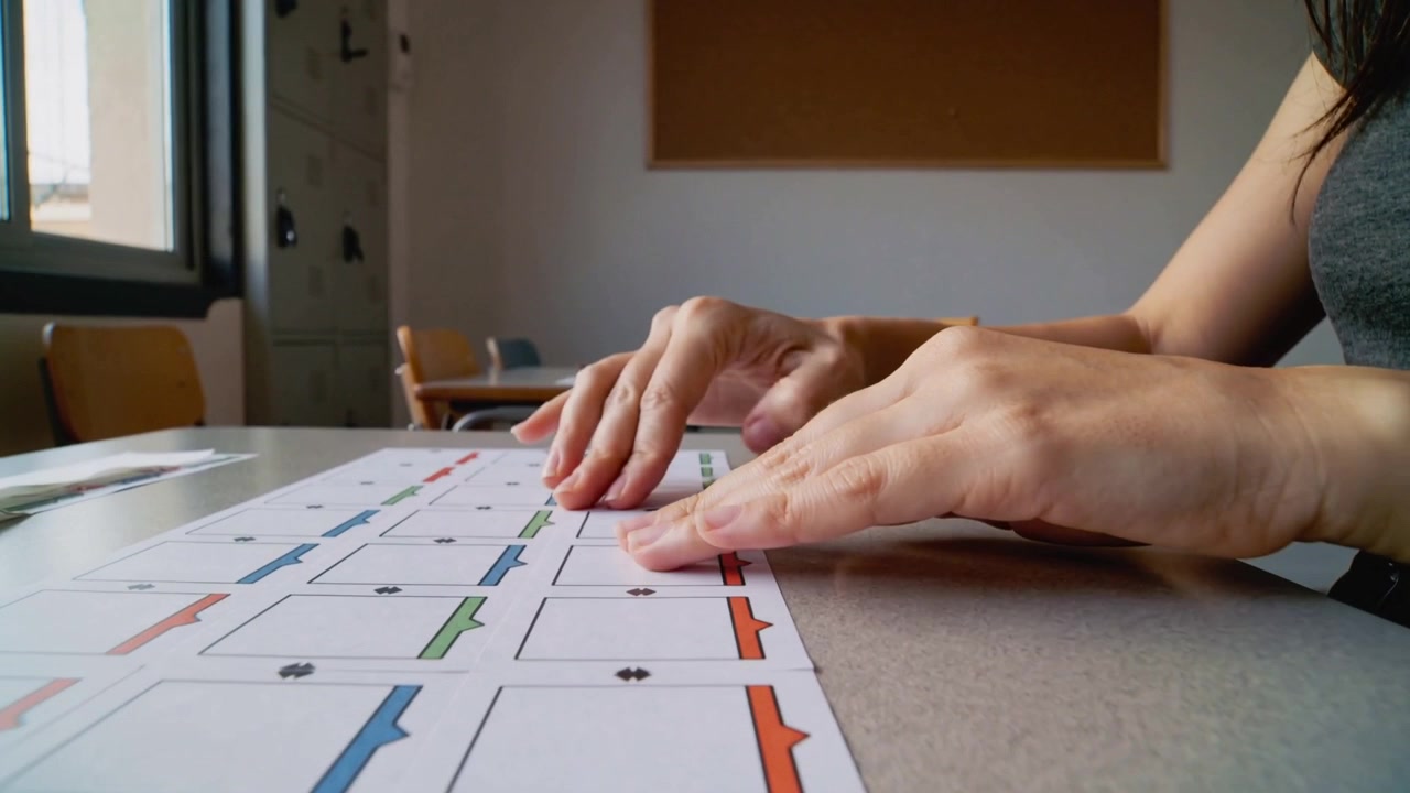 Picking up card, woman in sleeveless-top aligning cards on grid-sheet at table by window for study