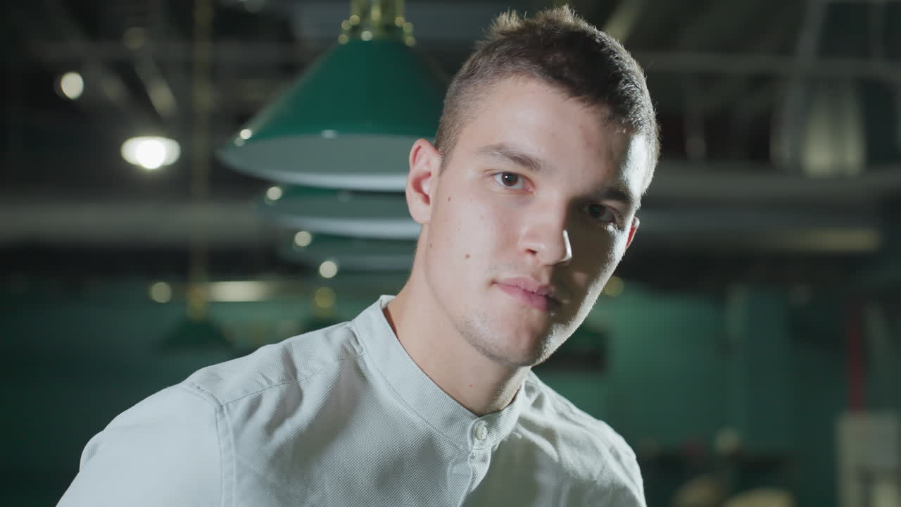 백색 셔츠를 입은 남자가 희미하게 조명된 빌리어드 홀에서 큐 스틱을 들고 있는 클로즈업. 오버헤드 조명은 그의 집중된 얼굴에 부드러운 그림자를 던집니다. 초록색 빌라 테이블과 매달린 조명은 대기 깊이를 만니다.