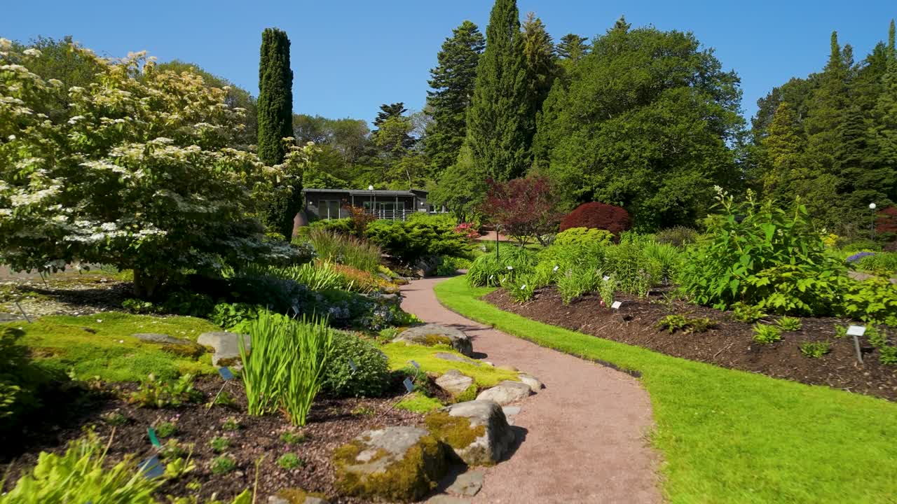 Płynny przelot dronem wzdłuż krętej ścieżki przez Ogród Botaniczny w Göteborgu, z bujnymi rabatami kwiatowymi, ogrodem skalnym i wysokimi drzewami iglastymi latem.