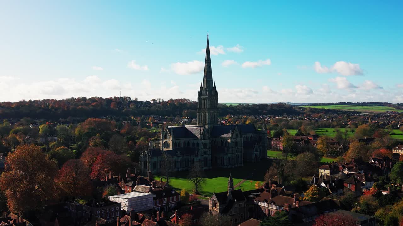 Riprese aeree con drone della Cattedrale di Salisbury con la sua alta guglia che si erge sopra edifici storici e alberi autunnali nella città di Salisbury, Inghilterra