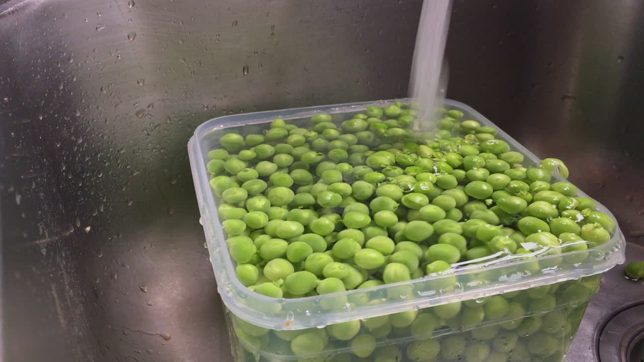 싱크 슬로우 모션에서 이녹스 콜랜더로 녹색 완두콩을 씻고 헹구는 여성의 영상