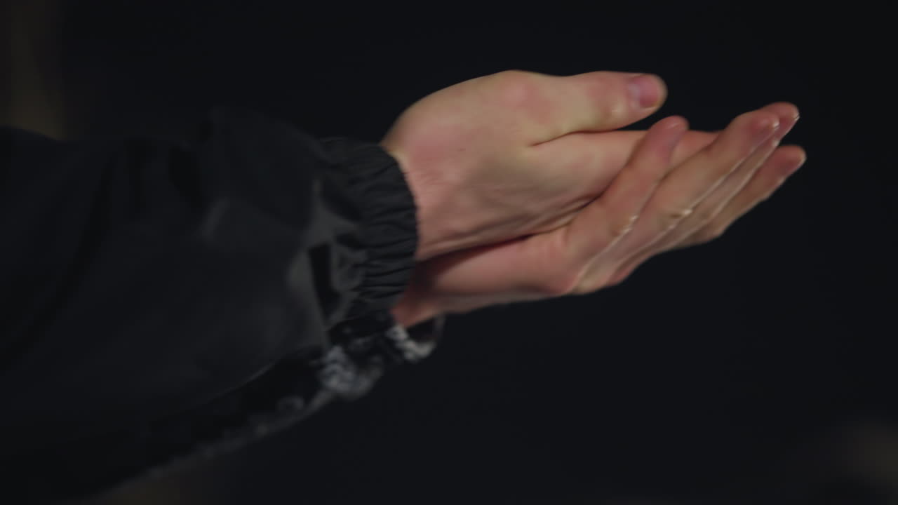 Close up of individual rubbing hands together for warmth during cold night outdoors, captured in low light with snowy background, conveying winter chill and natural response to freezing temperature