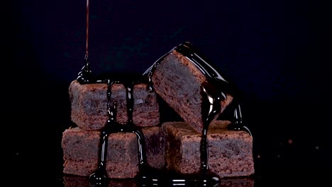 rich chocolate syrup pours slowly over a stack of brownie squares on a glossy black surface, captured in dramatic low-key lighting with a static camera