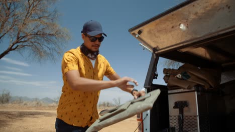 dynamic hand held tracking shot of a caucasian male tourist in africa as he takes out a grill bag with tongs from the back of a off-road vehicle and places it on a camping table.