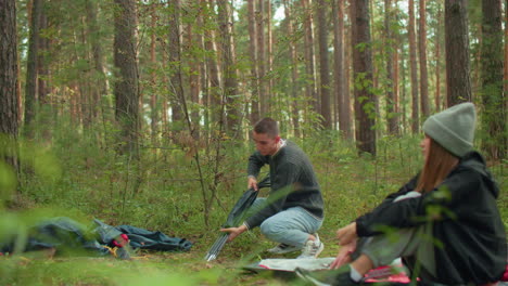 girl sitting on forest ground adjusting gray beanie while watching man in sweater crouch to unpack tent poles during camping setup surrounded by trees, scattered gear, and natural green foliage
