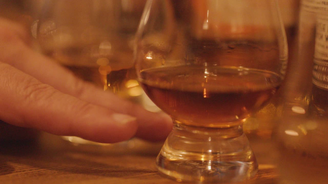 A hand gently rotates a whisky glass on a wooden table, surrounded by bottles and glasses, under soft, warm ambient lighting with shallow focus