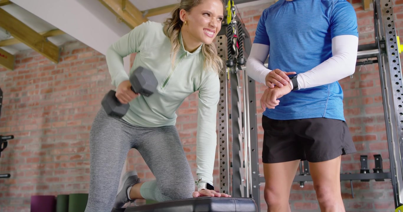 Træner tapper på smartwatch, angiver tempo, mens klienten udfører roning med sekskantede håndvægte på en stepbænk i fitnesscenteret.