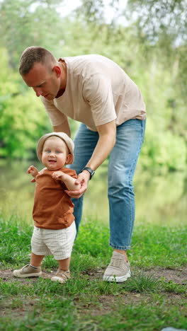 Loving dad leaning above his little baby learning to walk. Kid is going to drop but father catches him on time. Vertical video.