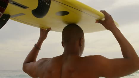 vista trasera de un surfista afroamericano que lleva una tabla de surf en la cabeza en la playa 4k