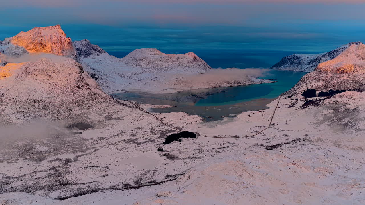 Norveç'in Lofoten bölgesinde, canlı bir kış gün batımı sırasında karla kaplı zirvelerin ve turkuaz bir fiyordun sinematik hava çekimi. Seyahat ve doğa belgeselleri için mükemmel.
